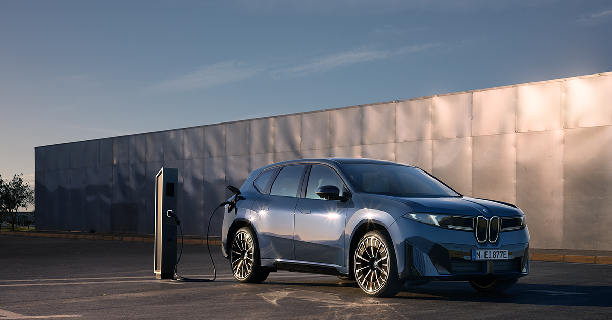 Das Bild zeigt den BMW iX3 beim Ladevorgang vor einer modernen Metallfassade. Das Design besticht durch die markante, vertikale Niere und eine cleane Linienführung in bläulichem Silber.
