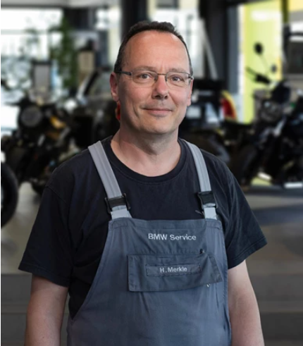 Ein Mann mit Brille und kurzen Haaren steht in einem Innenraum. Er trägt einen grauen BMW Service-Overall über einem schwarzen T-Shirt und blickt in die Kamera. Der Hintergrund ist unscharf, man kann jedoch Motorräder in einem Ausstellungsraum erkennen.