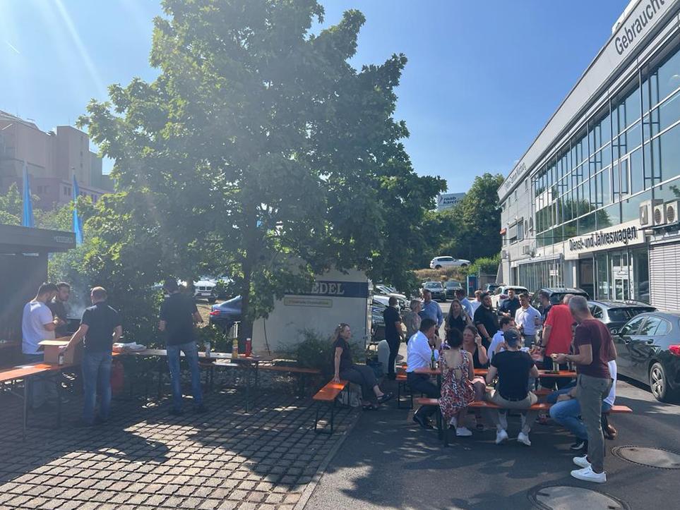 BMW Rhein Würzburg Grillen Menschen versammeln sich an Biertischen unter einem Baum vor einem Autohausgebäude.