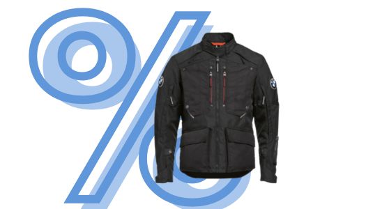 Im Fokus dieses Bildes steht eine schwarze, robuste Motorradjacke, die für das Touring- und Adventure-Segment gedacht ist. Die Jacke hat einen Stehkragen, mehrere Reißverschlusstaschen auf der Vorderseite und trägt kleine BMW-Logos an den Ärmeln. Rote Akzente finden sich an den Reißverschlüssen der Brusttaschen. Die Jacke wirkt sehr funktional und strapazierfähig. Auch hier überlagert das große, hellblau stilisierte Prozentzeichen (%) die gesamte Szene und bewirbt das Produkt im Rahmen eines Sales.