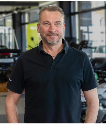 Das Bild zeigt einen Mann mittleren Alters mit einem grauen Bart und kurzen, grauen Haaren. Er blickt direkt in die Kamera und lächelt. Er trägt ein schwarzes Polo-Shirt. Der Hintergrund ist unscharf und zeigt einen Verkaufsraum für Motorräder.