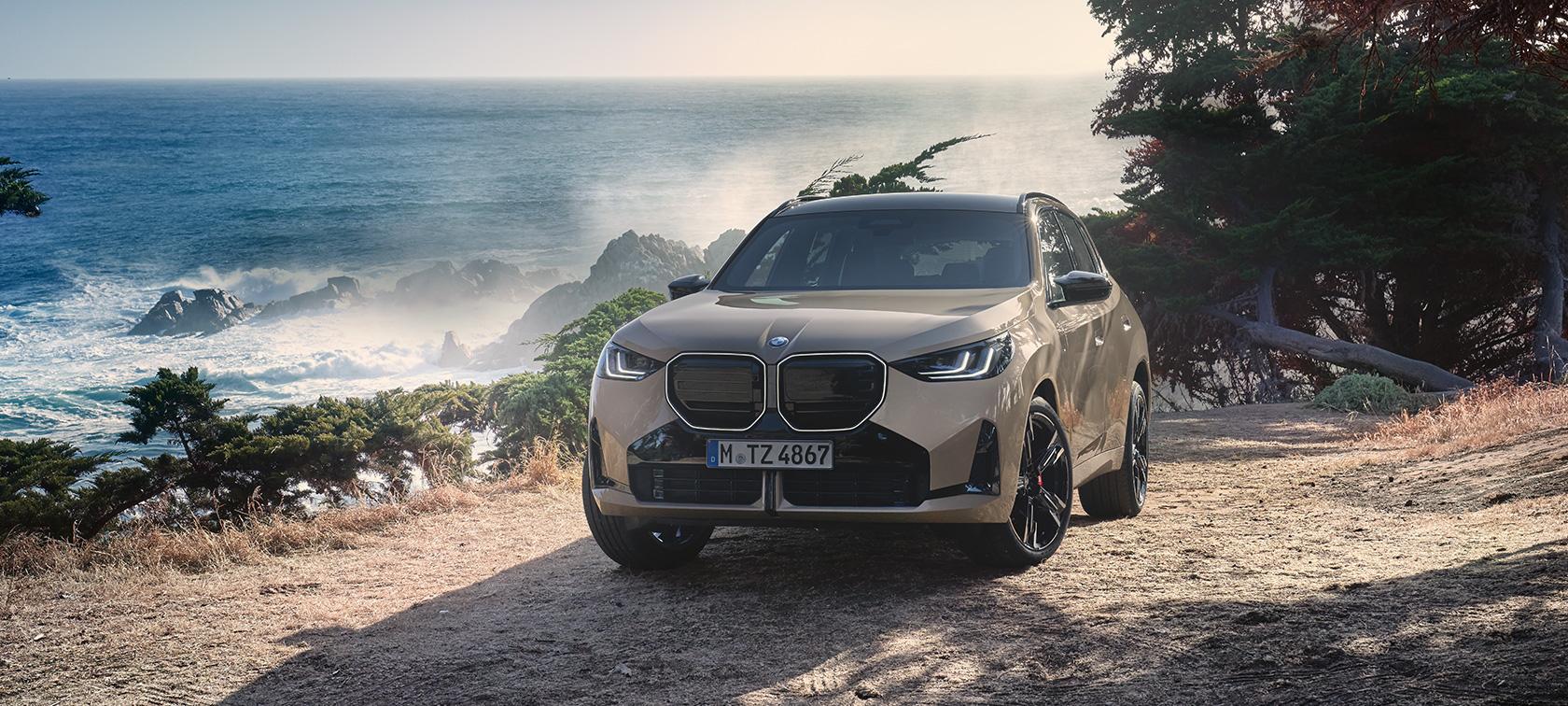 BMW X3 Rhein Gruppe Ein beigefarbener BMW X3 SUV steht auf einem Küstenweg mit Blick auf das Meer.