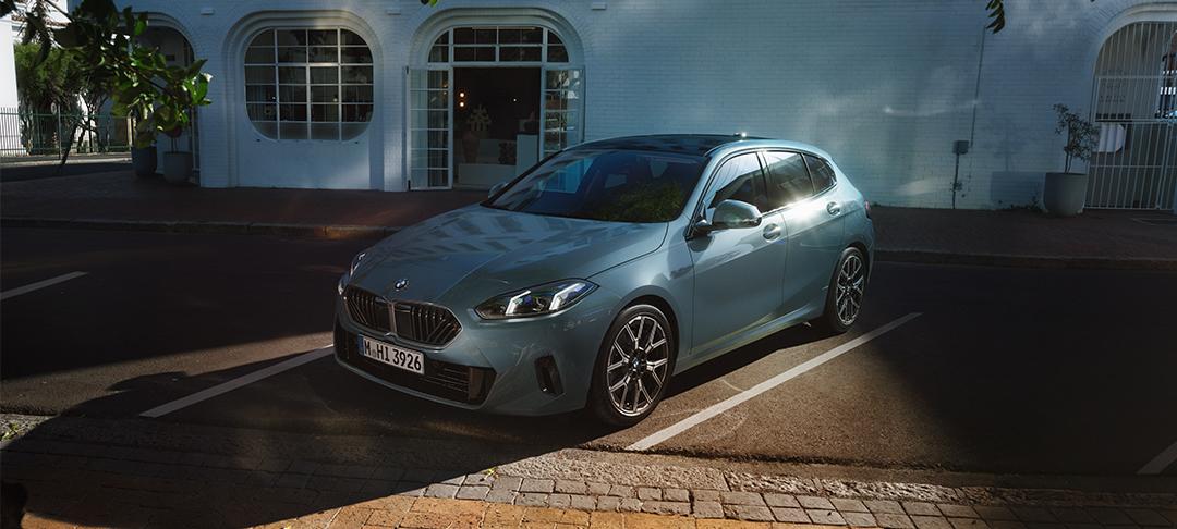 Das Bild zeigt einen blaugrauen BMW 1er (Modell 120), der auf einem markierten Parkplatz vor einem hellen Gebäude mit großen Rundbogenfenstern im Sonnenlicht parkt.