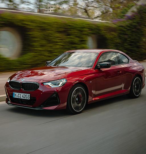 Das kompakte Coupé ist in einer dynamischen Vorbeifahrt eingefangen, wobei die Bewegungsunschärfe des Hintergrunds das Tempo unterstreicht. Die tiefrote Metallic-Lackierung bringt die muskulösen Formen und die lange Motorhaube besonders intensiv zur Geltung. Mit schwarzen Akzenten an der Niere und den Spiegelkappen wirkt das Fahrzeug extrem agil und fokussiert, was den puristischen Sportwagen-Charakter dieser Baureihe hervorhebt.
