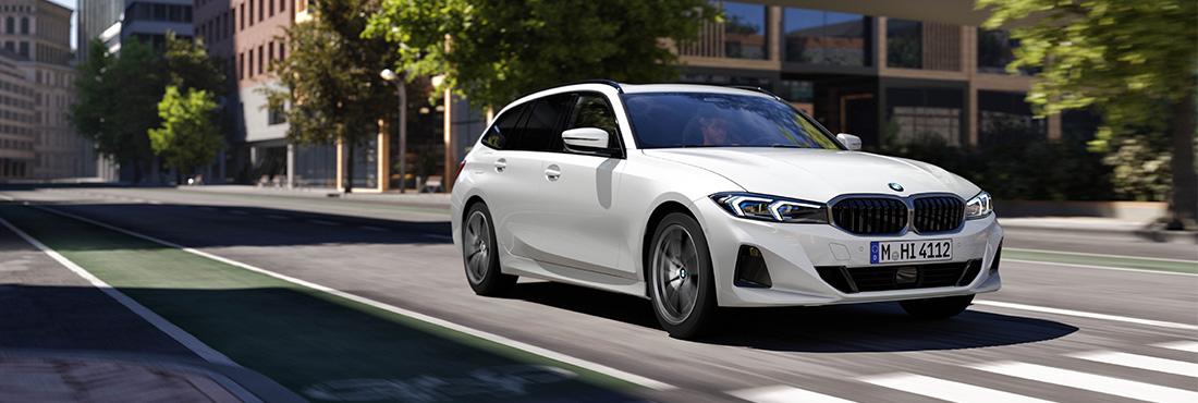 Das Bild zeigt einen weißen BMW 3er Touring, der dynamisch durch eine moderne Stadtkulisse fährt. Die Kombination aus der glänzenden Lackierung, den scharfen LED-Scheinwerfern und der leichten Bewegungsunschärfe auf dem Asphalt vermittelt einen sportlichen und hochwertigen Eindruck. Die Architektur im Hintergrund und das natürliche Sonnenlicht unterstreichen dabei den urbanen, eleganten Charakter des Fahrzeugs.