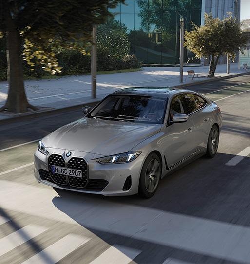 Es ist ein silbernes BMW 4er Gran Coupé zu sehen, das sich dynamisch auf einer innerstädtischen Straße bewegt. Das viertürige Coupé fällt besonders durch seine markante, große Niere an der Front auf und fährt an modernen Glasgebäuden und Bäumen vorbei.