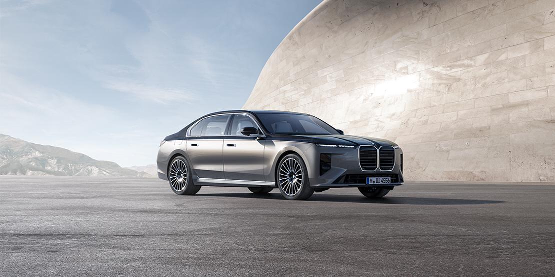 Eine zweifarbige BMW 7er Limousine in Silber und Schwarz parkt auf einer Betonfläche vor einer geschwungenen, modernen Betonwand unter blauem Himmel mit Bergen im Hintergrund.