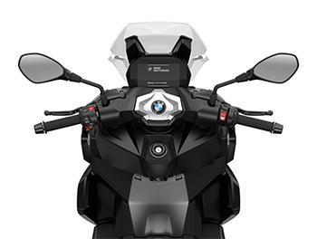 Das Bild zeigt die Ansicht direkt aus der Perspektive des Fahrers. Im Mittelpunkt steht das hochauflösende 6,5-Zoll-TFT-Farbdisplay, auf dem das BMW Motorrad Logo beim Starten zu sehen ist. Der Lenker wirkt aufgeräumt und hochwertig; links ist der bekannte Multi-Controller (Drehrad) zur Steuerung des Infotainments erkennbar. Das Bild vermittelt einen Eindruck von der Ergonomie und der modernen Konnektivität, die dem Fahrer Zugriff auf Navigation, Telefonie und Musik während der Fahrt ermöglicht.