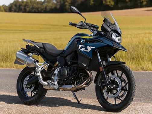  Auf dem Bild ist eine BMW F 800 GS abgebildet, ein mittelgroßes, dunkelblaues und gelbes Motorrad, das auf einer asphaltierten Straße steht.  Das Motorrad ist in einem hellen, sonnigen Licht und steht seitlich zur Kamera. Der Hintergrund ist eine grüne Wiese mit einigen gelben Blumen. Die Straße biegt leicht nach links ab und verläuft den Hang hinauf.