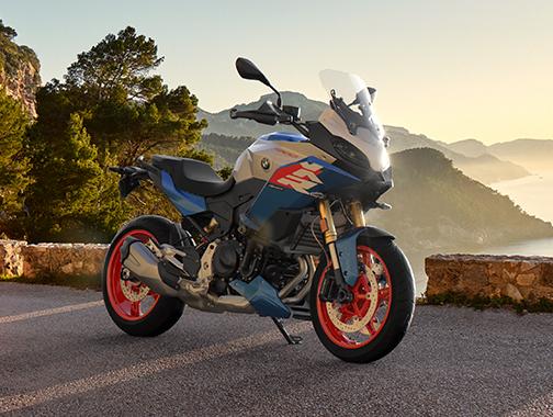  Auf dem Bild ist eine BMW F 900 XR abgebildet, ein mittelgroßes, blau-weißes und rotes Motorrad, das auf einer Straße mit Bergblick steht.  Das Motorrad ist in einem warmen, sonnigen Licht und steht seitlich zur Kamera. Es hat rote Räder, und die Karosserie ist in einem hellen Weiß und einem kräftigen Blau gehalten. Im Hintergrund befindet sich eine bergige Landschaft bei Sonnenuntergang. Das Motorrad ist sauber und glänzt, als wäre es brandneu.