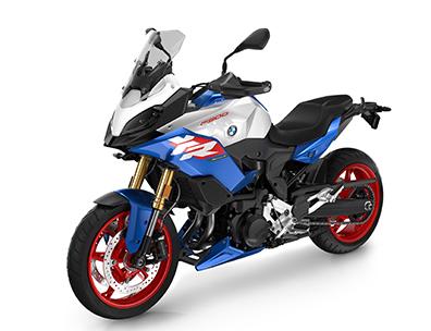  Auf dem Bild ist eine BMW F 900 XR abgebildet, ein mittelgroßes, blau-weißes und rotes Motorrad, das auf einem weißen Hintergrund steht.  Das Motorrad ist in einem hellen, sonnigen Licht und steht seitlich zur Kamera. Es hat rote Räder, und die Karosserie ist in einem hellen Weiß und einem kräftigen Blau gehalten. Es ist sauber und glänzt, als wäre es brandneu.