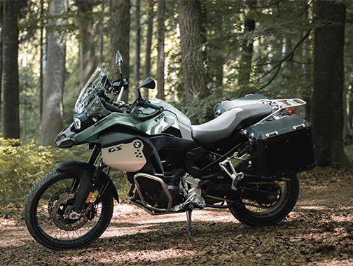  Auf dem Bild ist eine BMW F 900 GS Adventure abgebildet, ein großes, grünes, graues und schwarzes Motorrad, das in einem Wald steht.  Das Motorrad ist in einem hellen, sonnigen Licht und steht seitlich zur Kamera auf einem Boden mit Laub und Zweigen. Im Hintergrund befinden sich hohe, schlanke Bäume, die einen lichten Wald bilden. Das Motorrad ist sauber und glänzt, als wäre es brandneu.