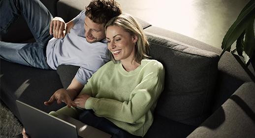 Auf diesem Bild ist das Paar in einer privateren, gemütlichen Umgebung zu sehen. Sie liegen entspannt auf einem großen, dunkelgrauen Sofa. Die Frau im Vordergrund trägt einen hellgrünen Pullover und blickt lächelnd auf einen Laptop, der auf ihrem Schoß steht. Der Mann liegt leicht versetzt hinter ihr, stützt sich auf seinen Ellenbogen und schaut ebenfalls interessiert auf den Bildschirm. Das sanfte Licht von oben rechts erzeugt eine wohnliche Stimmung und symbolisiert die Bequemlichkeit, ein Auto einfach von zu Hause aus zu konfigurieren oder zu bestellen.