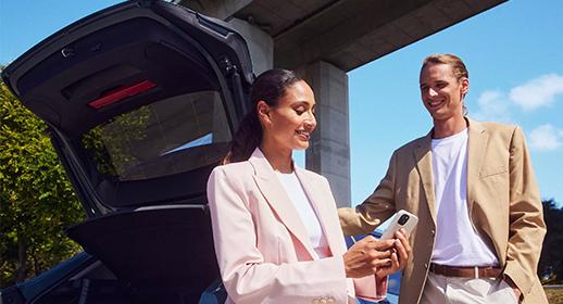 Das Foto zeigt eine Frau in einem hellrosa Business-Outfit und einen Mann in einem beigen Sakko, die draußen hinter einem Auto stehen. Die Heckklappe des Wagens ist weit geöffnet. Die Frau hält ein Smartphone in beiden Händen und blickt konzentriert auf das Display, während der Mann neben ihr steht und ebenfalls lächelnd auf das Handy schaut. Im Hintergrund ist eine moderne Betonbrücke und ein strahlend blauer Himmel zu sehen. Das Bild vermittelt Mobilität und die digitale Verwaltung von Finanzierungsthemen im Alltag.