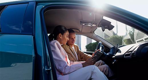 Hier sieht man ein Paar im Innenraum des Fahrzeugs. Die Frau sitzt auf dem Fahrersitz, der Mann auf dem Beifahrersitz. Die Fahrertür ist weit geöffnet, und helles Sonnenlicht fällt von oben in den Wagen. Gemeinsam blicken sie auf ein Tablet oder das große Display im Armaturenbrett des Autos. Sie scheinen sich mit den digitalen Funktionen oder zusätzlichen Services des Fahrzeugs vertraut zu machen. Die Perspektive von draußen durch die offene Tür lässt den Betrachter direkt am Geschehen teilhaben.