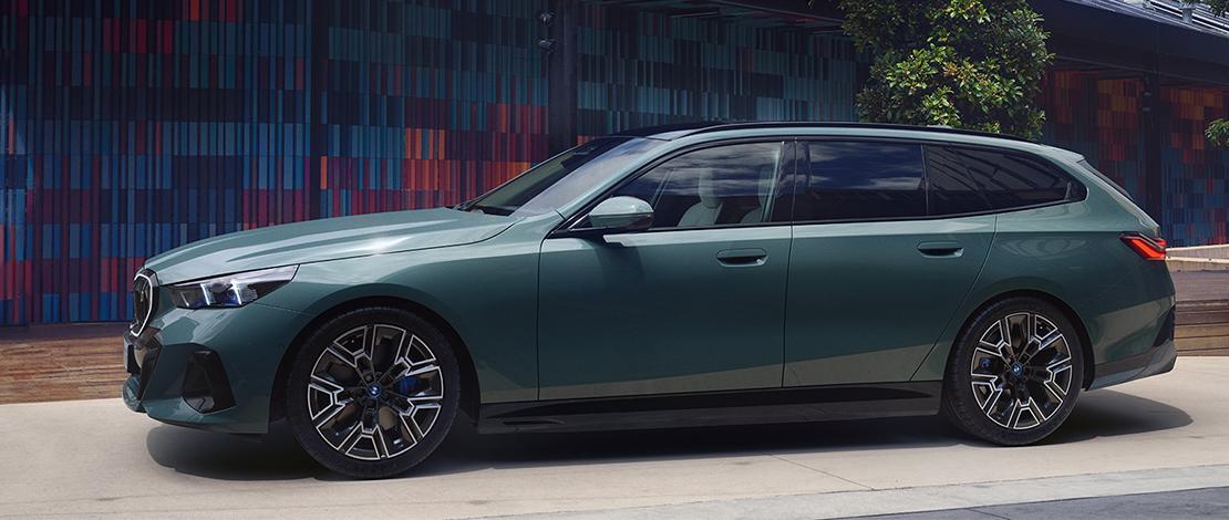Ein blassgrüner BMW i5 Touring, eine Kombi-Limousine, ist im Profil auf einem gepflasterten Parkplatz abgebildet. Das Auto hat schwarze Felgen mit blauen Bremssätteln und getönte Scheiben. Im Hintergrund ist ein Gebäude mit einer farbenfrohen, modernen Fassade zu sehen, die aus verschiedenen rechteckigen Abschnitten in Blau-, Orange- und Brauntönen besteht.