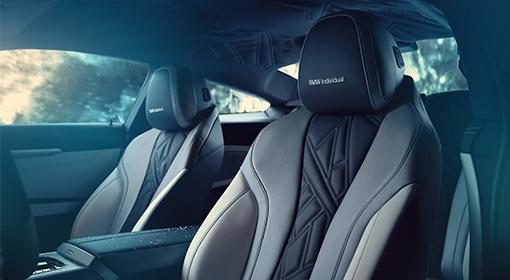 Nahaufnahme der sportlichen Vordersitze eines BMW-Fahrzeugs. Die Sitze sind zweifarbig in Dunkelgrau und hellem Beige gehalten, mit einem markanten, rautenförmigen Steppmuster auf der mittleren Rückenlehne. Auf den Kopfstützen ist "BMW Individual" eingestickt.