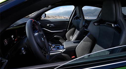 Innenansicht eines sportlichen BMW M-Fahrzeugs, fokussiert auf den Fahrerbereich. Man sieht das Lenkrad mit roten Akzenten, das digitale Cockpit und die dunklen Sportsitze. Durch das Seitenfenster ist eine Landschaft mit Bergen und Wasser zu erkennen.