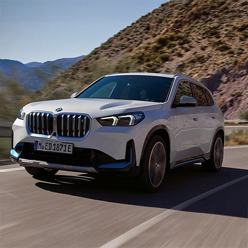Ein weißer BMW iX1 SUV fährt dynamisch auf einer kurvigen Bergstraße entlang. Im Hintergrund sind felsige Berghänge und ein blauer Himmel zu sehen