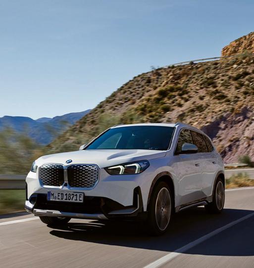 Dieses Foto zeigt einen weißen BMW iX1 xDrive30, ein kompaktes SUV, in einer dynamischen Fahrsituation auf einer Bergstraße.  Das Auto ist von vorne leicht seitlich aufgenommen und wirkt, als würde es schnell über eine asphaltierte Straße fahren. Die Frontpartie ist dominant, mit der typischen BMW-Niere, die hier geschlossen und gitterartig (charakteristisch für die i-Modelle) gestaltet ist.  Im Hintergrund erheben sich hohe, trockene Berge mit rötlich-brauner Erde unter einem klaren, blauen Himmel. Es ist ein sonniger Tag. Auf dem Nummernschild ist 'M ED 1871 E' zu lesen.