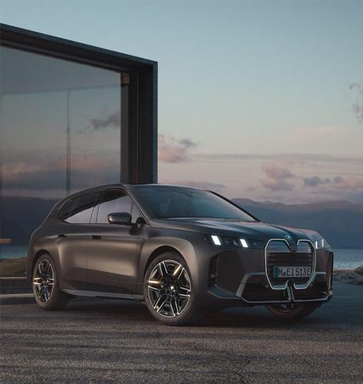 Das Bild zeigt einen dunkelgrauen oder schwarzen BMW iX M70 xDrive im Dreiviertelprofil von vorne rechts. Es ist ein großes, futuristisch aussehendes Elektro-SUV.  Das Fahrzeug steht auf einer asphaltierten Fläche neben einem modernen Gebäude mit großen Glasflächen, wahrscheinlich in der Abenddämmerung oder bei Sonnenuntergang. Die Front ist durch die große, aufrechte, geschlossene Nierenform und die schmalen, hell leuchtenden Scheinwerfer sehr präsent.  Im Hintergrund ist eine Küstenlandschaft mit einem See oder Meer zu sehen, darüber ein Himmel mit sanften Wolken und tiefstehender Sonne. Das Nummernschild ist 'M EI 513 E'. Das gesamte Bild hat eine sehr gedämpfte und hochwertige Ästhetik.