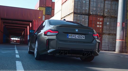Heckansicht eines grauen BMW M2, der auf einer Straße zwischen hohen Stapeln roter und grauer Schiffscontainer fährt.