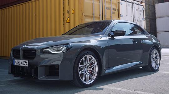 Ein sportlicher, dunkelgrauer BMW M2 steht schräg vor hohen, gestapelten gelben und weißen Schiffscontainern.