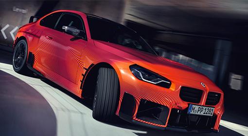 Ein roter BMW M2, ausgestattet mit M Performance Parts, fährt mit hoher Geschwindigkeit um eine Kurve. Das Auto ist mit einem auffälligen orange-schwarzen gestreiften Muster verziert und trägt das Kennzeichen "M PP 1281".