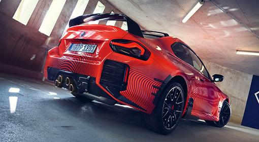 Heckansicht eines roten BMW M2 mit schwarzen M Performance Parts, darunter ein großer Heckflügel und ein markanter Diffusor mit vier goldenen Auspuffrohren. Das Fahrzeug steht schräg in einer Parkgarage mit seitlich einfallendem Licht.