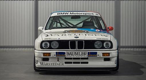 Frontansicht eines weißen BMW M3 E30 Rennwagens mit blauen und roten BMW Motorsport Streifen und Sponsorenlogos.