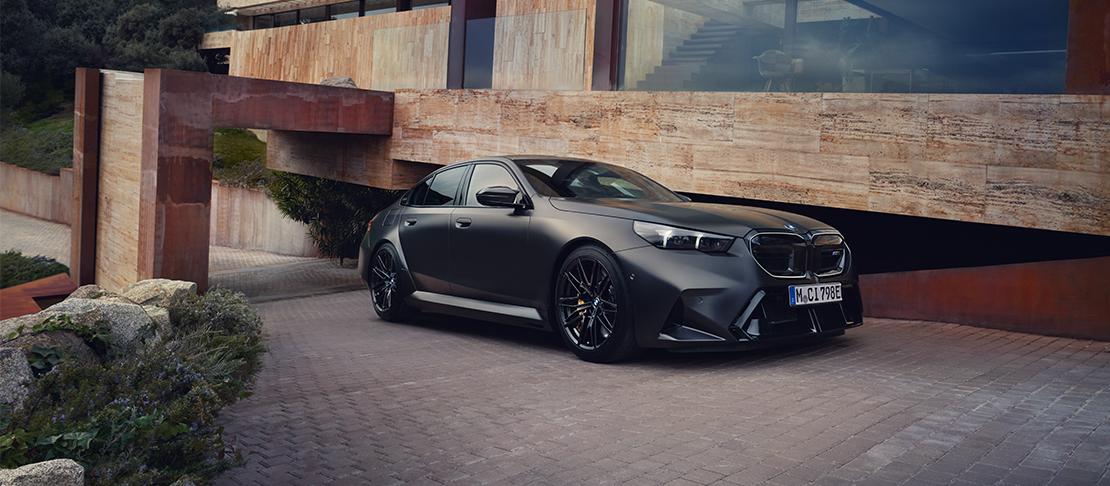 Eine mattschwarze BMW M5 Limousine präsentiert sich kraftvoll vor einer modernen Luxusvilla aus Naturstein. Das Zusammenspiel aus exklusiver Architektur, sportlichen Details wie gelben Bremssätteln und der eleganten, diffusen Lichtstimmung unterstreicht den High-End-Anspruch des Hybrid-Sportlers.