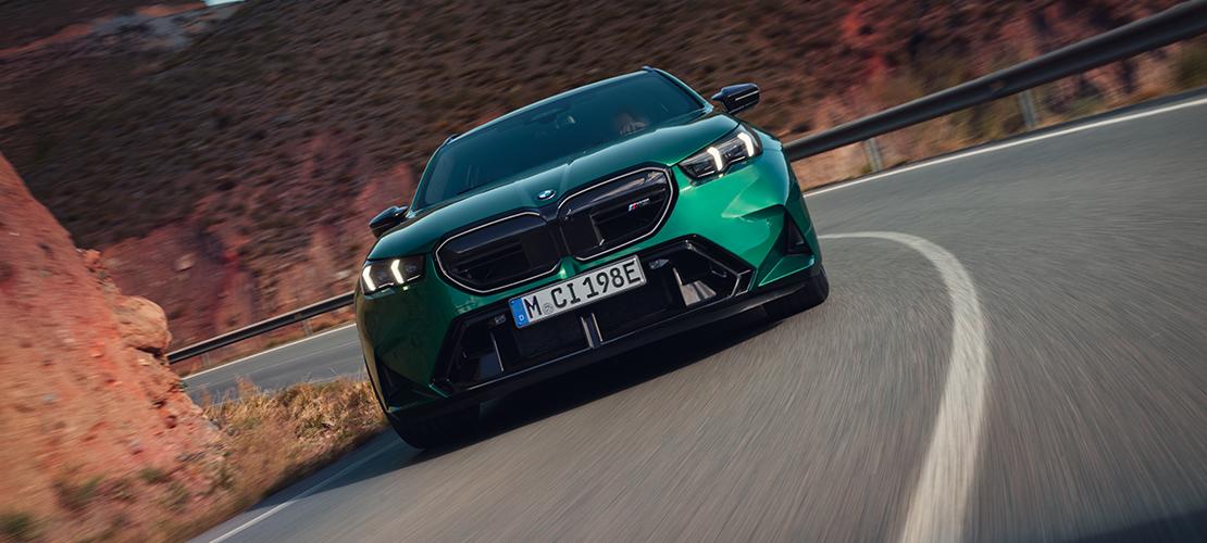 Auf dem Bild ist ein grüner BMW M5 Touring in Bewegung zu sehen. Das Auto fährt auf einer kurvigen Bergstraße. Im Hintergrund sind felsige, rötliche Felsen erkennbar. Das Licht ist eher gedämpft, und das Bild vermittelt eine sportliche, dynamische Atmosphäre.