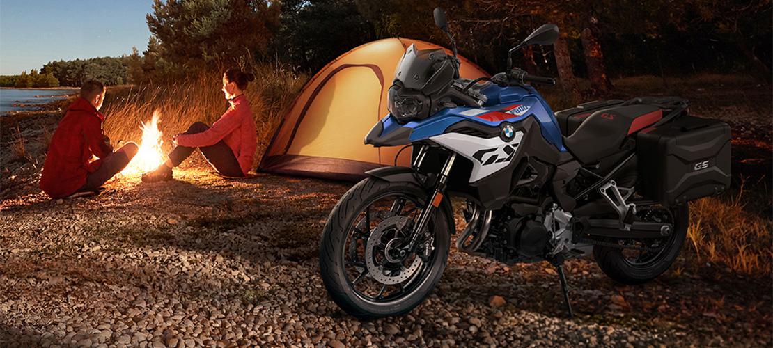 BMW F 800 GS Motorrad am Seeufer mit Campern am Lagerfeuer bei Sonnenuntergang.