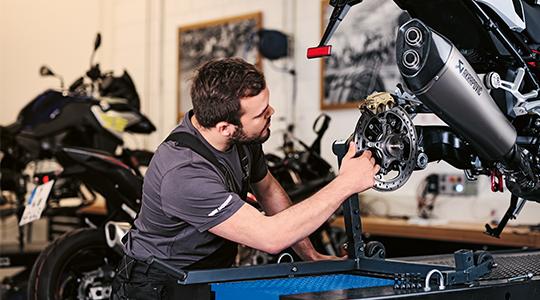 Ein Mechaniker prüft die hintere Bremsanlage einer Maschine mit Akrapovič-Auspuff, die auf einer Hebebühne aufgebockt ist.
