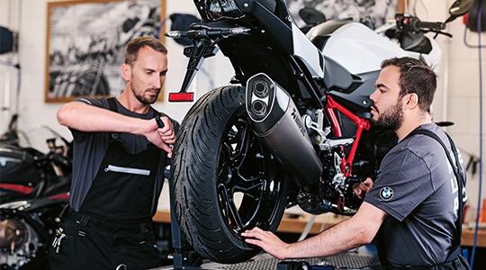 Zwei Mechaniker arbeiten gemeinsam am Hinterrad eines Motorrads; einer fixiert das Rad, während der andere Montagearbeiten vornimmt.