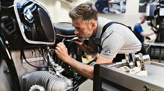 Ein erfahrener Mechaniker prüft konzentriert die Details eines klassischen Boxermotors (Oldtimer/Heritage-Modell), während im Vordergrund Motorkomponenten auf einem Werkstattwagen liegen.