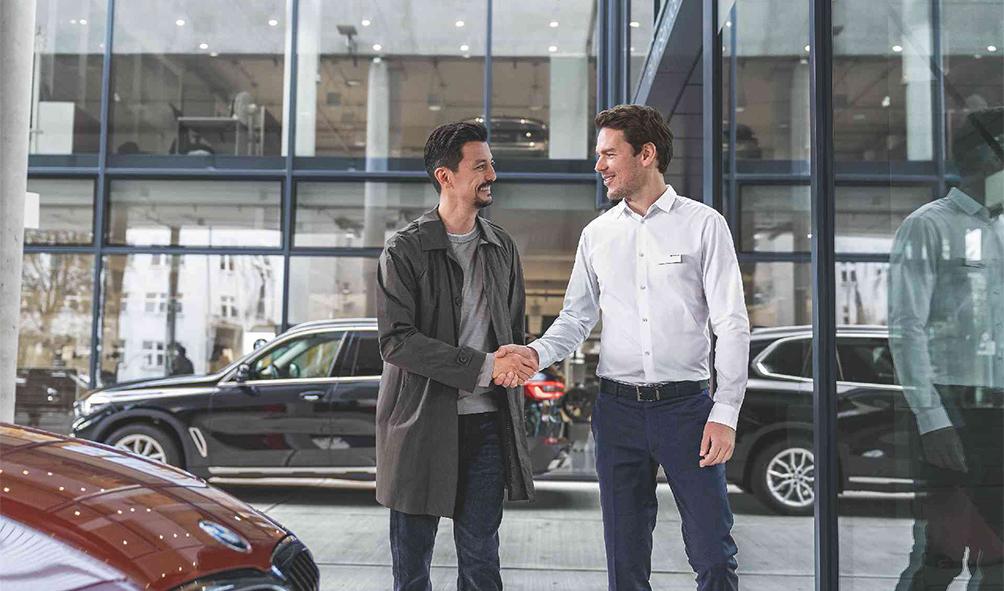 Das Bild zeigt zwei Männer in einem Autohaus, die sich lächelnd die Hand geben – vermutlich ein Kunde und ein Autoverkäufer nach einem gelungenen Gespräch, vor der Kulisse von ausgetellten Fahrzeugen und einer Glasfront.