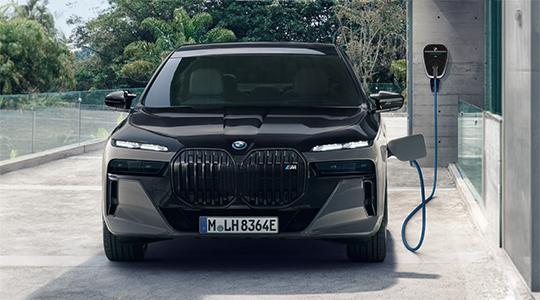 Das Bild zeigt die Frontansicht einer schwarzen BMW 7er Limousine als Plug-in-Hybrid. Das Auto ist in einer Einfahrt geparkt und wird an einer grauen Wallbox aufgeladen. Die Frontleuchten sind eingeschaltet und die Nieren sind im Vordergrund. Im Hintergrund sind grüne Pflanzen zu sehen.