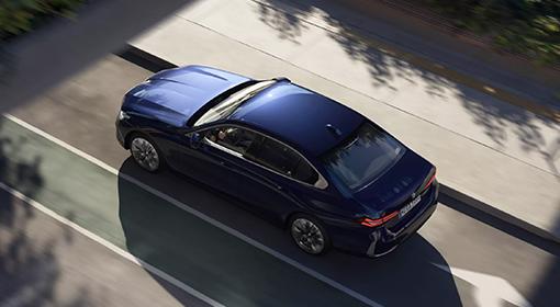 Das Bild zeigt eine dunkelblaue BMW 5er Limousine Plug-in-Hybrid, die auf einer Straße fährt. Die Aufnahme ist von schräg oben gemacht, sodass das Dach und die Motorhaube gut zu sehen sind. Auf der Straße sind helle Linien und im Hintergrund sind die Schatten von Bäumen zu sehen.