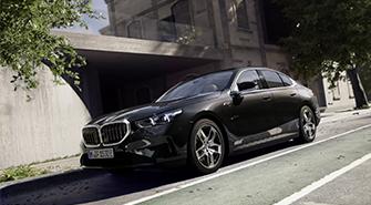 Das Bild zeigt die Seitenansicht eines dunklen BMW 5er Plug-in-Hybrid, der auf einer Straße geparkt ist. Das Auto steht leicht schräg mit dem vorderen Teil nach links. Im Hintergrund sind ein Gebäude und Bäume zu sehen. Die Frontleuchten des Autos sind eingeschaltet.