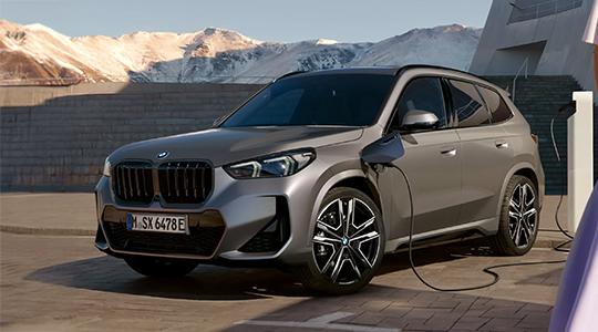 Das Bild zeigt die Front eines grauen BMW X1 Plug-in-Hybrid. Das Auto ist draußen auf einem gepflasterten Platz geparkt. Es wird an einer Ladesäule aufgeladen, die sich auf der rechten Seite befindet. Im Hintergrund sind Berge mit Schnee zu sehen.