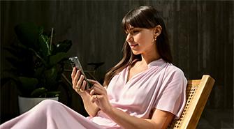 Das Bild zeigt eine junge Frau in einem hellrosa Kleid, die auf einem Stuhl sitzt. Sie schaut lächelnd auf ihr Smartphone, das sie mit beiden Händen hält. Im Hintergrund ist eine dunkle Wand und eine große grüne Pflanze zu sehen. Die Szene ist von Sonnenlicht beleuchtet.