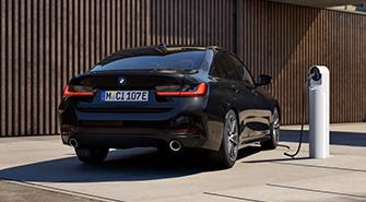 Das Bild zeigt die Heckansicht eines schwarzen BMW 3er Plug-in-Hybrid. Das Auto steht draußen vor einer modernen Hausfassade mit Holzverkleidung. Die Rückleuchten sind an. Rechts neben dem Auto befindet sich eine Ladesäule mit einem angeschlossenen Kabel, das in das Auto führt.