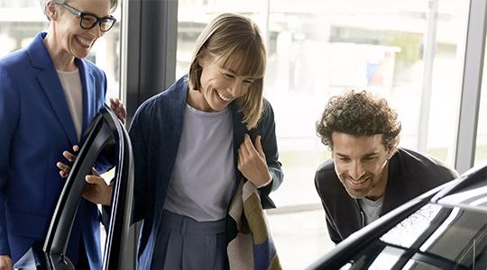 Das Bild zeigt drei Personen in einem Autohaus, die sichtlich fröhlich sind. Sie stehen um ein Auto herum, das nur teilweise zu sehen ist. Eine Frau in einem blauen Blazer, die eine Brille trägt, lacht und schaut in den Innenraum des Autos. Eine andere Frau mit kurzen Haaren in einer blauen Jacke schaut ebenfalls lächelnd in den Innenraum des Wagens. Ein Mann mit lockigen Haaren beugt sich ebenfalls lachend über die Motorhaube des Autos. Die Atmosphäre wirkt sehr positiv und entspannt.