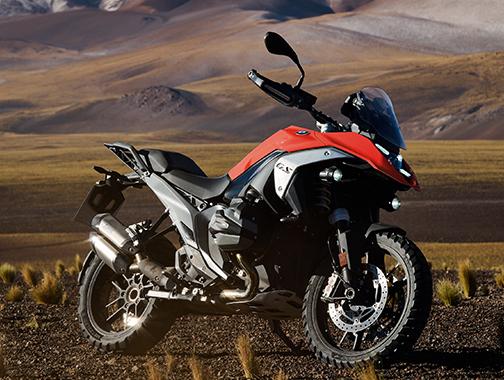  Auf dem Bild ist eine BMW R 1300 GS abgebildet, ein großes, rotes und schwarzes Motorrad, das in einer Wüsten- oder Berglandschaft steht.  Das Motorrad ist in einem hellen, sonnigen Licht und steht seitlich zur Kamera auf einem sandigen Boden. Der Hintergrund ist eine bergige Wüste mit ockerfarbenem Sand und vereinzelten, braunen Grasbüscheln. Es ist sauber und glänzt, als wäre es brandneu.
