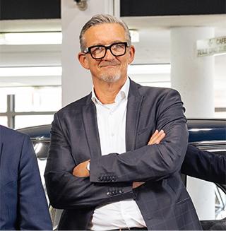 Lächelnder Mann mit Brille und Anzug, Arme verschränkt, in einem Autohaus an ein Auto gelehnt.
