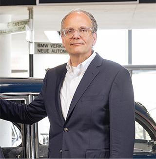 Älterer Herr mit Brille und Anzug steht lächelnd vor einem Oldtimer in einem BMW Autohaus.