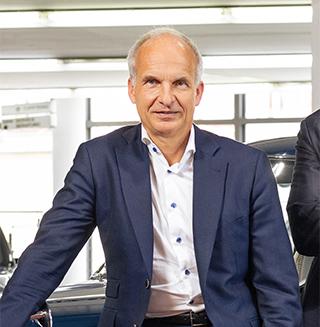 Ein lächelnder Mann in blauem Anzug lehnt in einem Autohaus an einem blauen Oldtimer.e