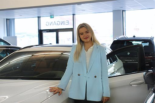 Eine junge, lächelnde Frau lehnt an einem Auto in einem BMW Autohaus.
