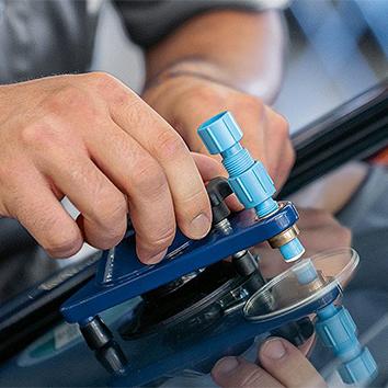 Das Bild zeigt eine Detailaufnahme von Händen bei einer Steinschlagreparatur an einer Fahrzeugscheibe, bei der ein spezialisiertes blaues Werkzeug mit Injektor und Saugnapf präzise auf der Glasscheibe fixiert wird.